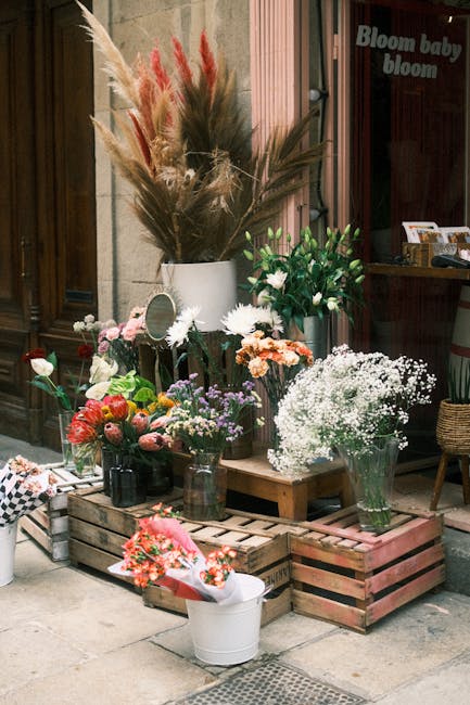A display of various fresh flowers arranged in vases and jars outdoors in front of a wooden storefront, featuring a mix of colorful blooms such as pink roses, white daisies, baby's breath, and lavende