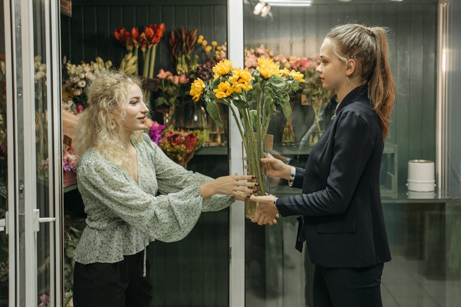A florist shop with a display of various floral arrangements and bouquets, featuring vibrant yellow, pink, red, and purple flowers arranged in vases and bouquets. In the foreground, a woman with blond