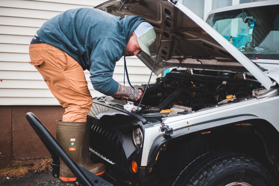 A man wearing a green cap, blue sweatshirt, khaki pants, and green rubber boots is leaning over the open hood of a silver off-road vehicle, inspecting or working on the engine. The vehicle is parked o