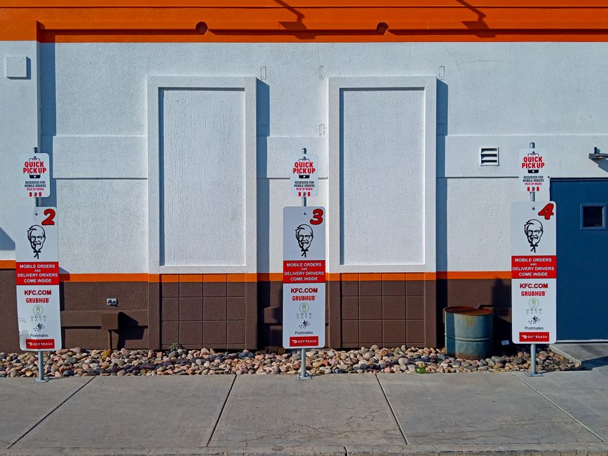 A plain white wall of a fast-food restaurant with four numbered KFC quick pickup signs positioned on the sidewalk in front. Each sign features the KFC logo with Colonel Sanders' image, red accents, an