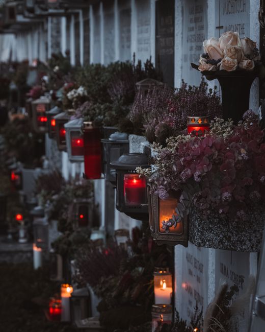 A row of memorial graves adorned with fresh floral arrangements and softly glowing red and orange lanterns. The flowers include pale pink roses, white blossoms, and various purple and burgundy blooms,