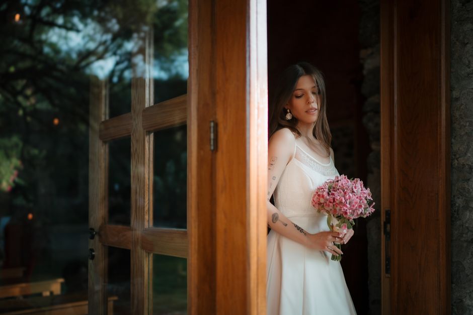 A young woman in a white wedding dress with lace accents, holding a floral bouquet of pink hydrangeas, stands partially visible through an open wooden door framed by stone walls, with natural light il