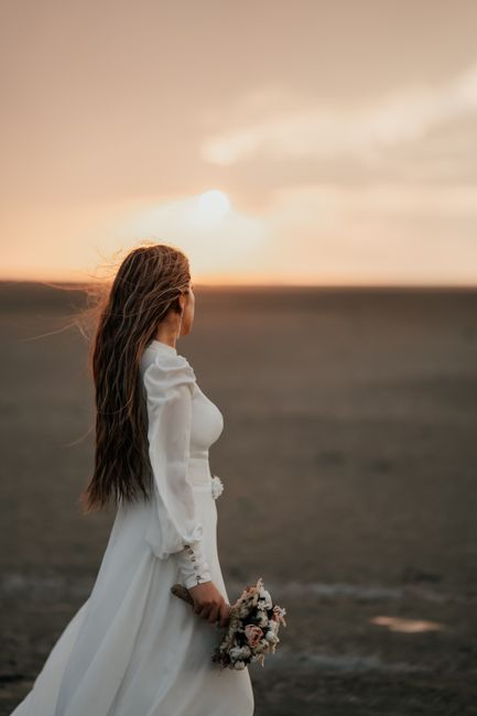A bride dressed in a white wedding gown with long sleeves and lace details, holding a small bouquet of mixed pastel-colored flowers including roses and greenery, standing on a sandy beach during sunse